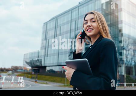 Una giovane donna vestita con una tuta formale si erge fuori da un edificio di uffici contemporaneo, sorridendo mentre parla al telefono e tiene in mano un tablet, consiglia Foto Stock