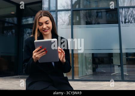Una donna è seduta su una panchina di fronte a un edificio contemporaneo, utilizza un tablet e sorride mentre interagisce con i contenuti sullo schermo, godendosi Foto Stock