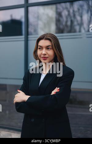 Una giovane donna sta in piedi con le braccia incrociate di fronte a un edificio contemporaneo. Sorride leggermente, vestita con un blazer nero con un top bianco, Foto Stock