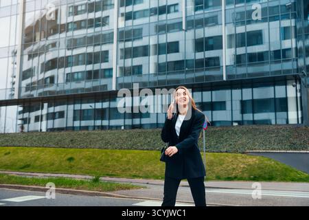 Una donna cammina con sicurezza sul marciapiede mentre parla sul suo smartphone. È vestita con abiti professionali e il moderno edificio per uffici Foto Stock