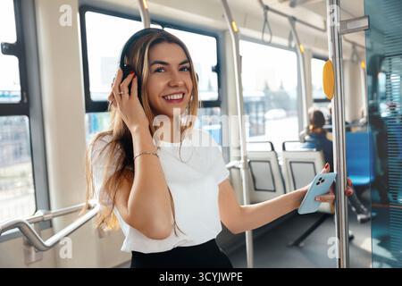 Una giovane donna sta su un veicolo di trasporto pubblico, sorridendo mentre ascolta la musica attraverso le cuffie. Possiede uno smartphone e si diverte a spostarsi. Foto Stock