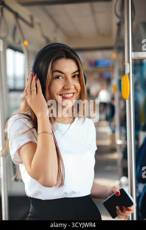 Una giovane donna con i capelli lunghi sorride mentre ascolta la musica con le cuffie all'interno di un autobus cittadino. Possiede uno smartphone e si diverte a viaggiare in mezzo a fel Foto Stock
