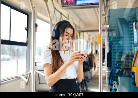 Una giovane donna con i capelli lunghi ama ascoltare la musica con le cuffie mentre sorride al suo smartphone all'interno di un veicolo di trasporto pubblico durante le ore diurne. Foto Stock