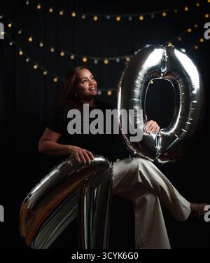 Una donna ha in mano un palloncino d'argento che dice 20. Lei è seduta per terra. Ci sono altre bollature sullo sfondo Foto Stock