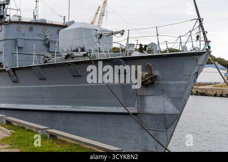 Primo piano della prua e della sovrastruttura anteriore di una grande, vecchia nave militare o pattuglia grigia ormeggiata in un porto, con un'ancora pesante visibile. Foto Stock