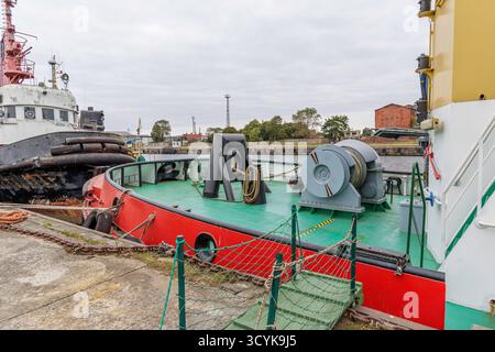 Un ampio ed esteso paesaggio di un porto industriale caratterizzato da due rimorchiatori arancioni e bianchi attraccati in primo piano, con una vasta, verde e lontana sponda Foto Stock