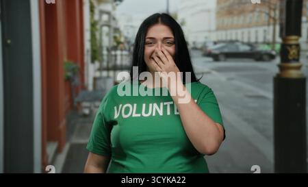 Donna sorridente che indossa una camicia verde volontaria in piedi per strada in città con una bocca che copre a mano l'energia giovanile e lo spirito della comunità all'aperto Foto Stock
