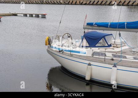 Primo piano della sezione anteriore di una barca a vela bianca con accenti blu, ancorata in un porto tranquillo con volante e ponte in legno visibile. Foto Stock
