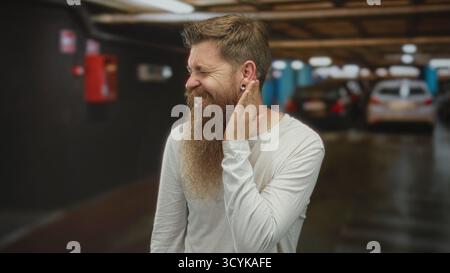 L'uomo con barba folta e camicia bianca informale tiene mano a mano nell'edificio pieno di auto sfocate che riflettono le luci sopra la testa; disagio. Foto Stock