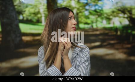 La donna stringe le mani sotto il mento e guarda con ansia al lato sotto il tettuccio verde illuminato dal sole nella foresta; preoccupazione. Foto Stock