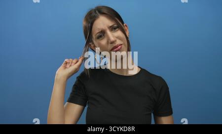 Una donna tiene una ciocca di capelli tra le dita in un ambiente blu da studio con una nebbia perplessa; incertezza. Foto Stock