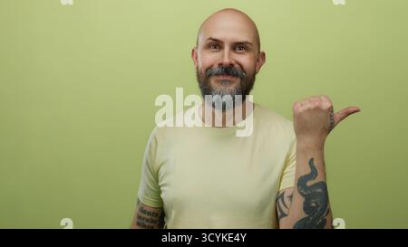 Uomo dalla barba calva con gesti tatuati con il pollice su sfondo isolato dalla parete gialla Foto Stock