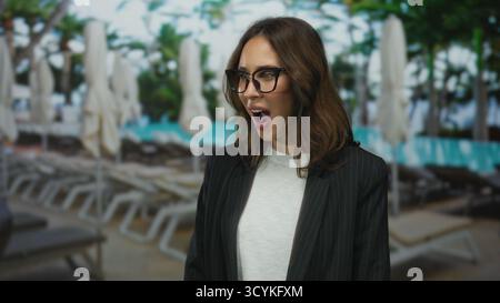 Donna che indossa occhiali e camicia bianca, sorpresa nella piscina esterna del resort di fronte a sdraio e ombrelloni, suggerendo svago in un ambiente alberghiero. Foto Stock