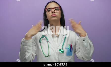 La dottoressa ispanica con uno stetoscopio si raffredda con le mani davanti a una parete viola, indossando occhiali e un'uniforme bianca, esprimendo calore Foto Stock