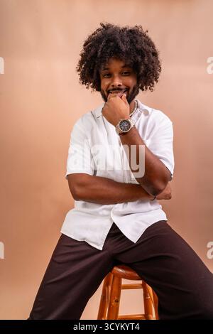 Giovane uomo con capelli ricci che posa felicemente su uno sgabello in uno studio Foto Stock