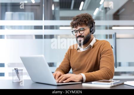 Uomo sorridente che indossa cuffie e occhiali mentre digita un laptop, fornisce assistenza clienti o si impegna a comunicare online da un ufficio moderno Foto Stock