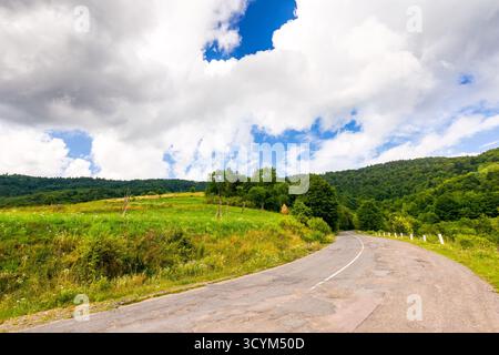 passo uzhaniano nel paesaggio montano dei carpazi. la vecchia strada di campagna asfaltata scende in discesa vicino alla foresta. scena rurale con campi e fienili in somme Foto Stock