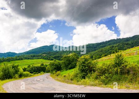 passo uzhaniano nel paesaggio montano dei carpazi. la vecchia strada di campagna asfaltata scende in discesa vicino alla foresta. scena rurale con campi e fienili in somme Foto Stock