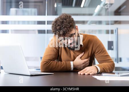 Giovane uomo d'affari che soffre di dolore acuto al petto mentre è seduto alla scrivania con un portatile, indicando potenziali problemi di salute legati allo stress o un'improvvisa emergenza medica Foto Stock