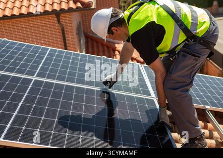 Un individuo che indossa un casco di sicurezza e un giubbotto riflettente lavora con attenzione all'installazione di pannelli solari su un tetto. I cieli azzurri offrono un clima ideale Foto Stock