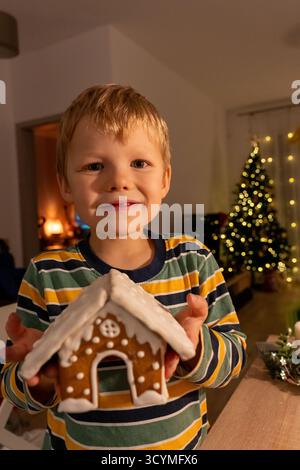 Felice biondo in maglione a righe che mostra orgogliosamente la casa di pan di zenzero fatta in casa con caminetto e luci dell'albero di Natale in un accogliente interno. Vacanze Foto Stock