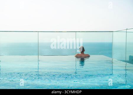 Uomo che si rilassa nella piscina a sfioro sul tetto con orizzonte oceano senza fine sotto il cielo limpido sull'isola tropicale di Phu Quoc, Vietnam. Gli uomini godono di vista aerea sul mare Foto Stock