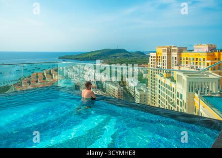 Uomo che si rilassa nella piscina a sfioro sul tetto con splendida vista panoramica dell'isola tropicale e degli edifici in stile mediterraneo nella città Sunset di Phu Quoc Foto Stock