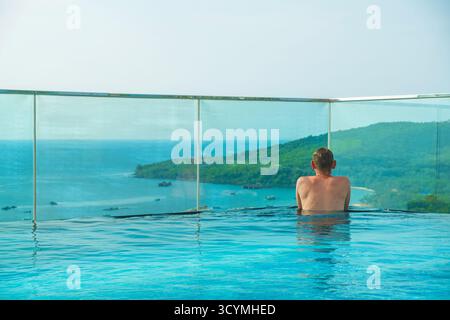 Uomo che si rilassa nella piscina a sfioro sul tetto con splendida vista panoramica dell'isola tropicale e degli edifici in stile mediterraneo nella città Sunset di Phu Quoc Foto Stock
