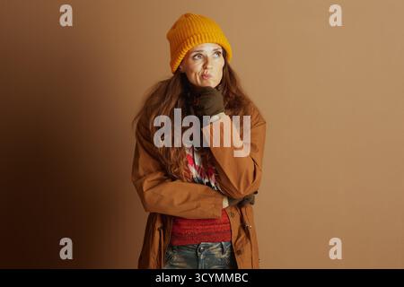 Una donna premurosa con un berretto giallo, un cappotto marrone e un maglione rosso poggia il mento sulla mano, guardando verso l'alto con un'espressione incerta contro un pla Foto Stock