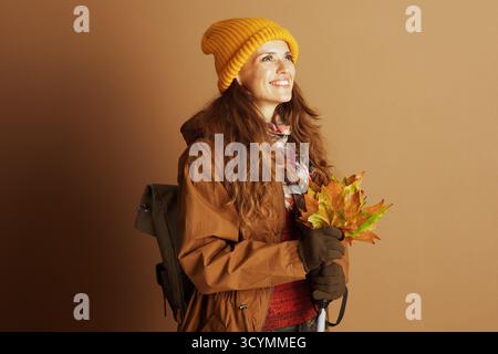 Una donna di mezza età, decorata con berretto giallo e giacca con cappuccio, sorride verso l'alto tenendo in mano un bouquet di foglie autunnali e bastone da passeggio. Il suo allegro Foto Stock
