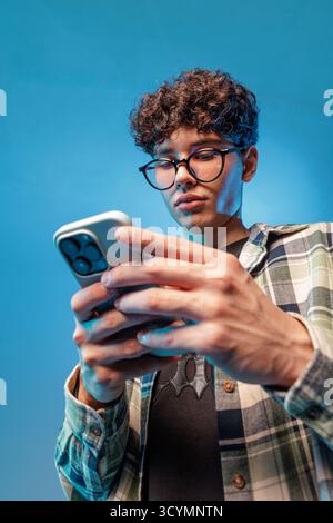 Teenager concentrato con occhiali utilizzando uno smartphone in studio, che illustra l'ora dello schermo, la luce blu, i messaggi di testo e le moderne abitudini digitali. Foto Stock