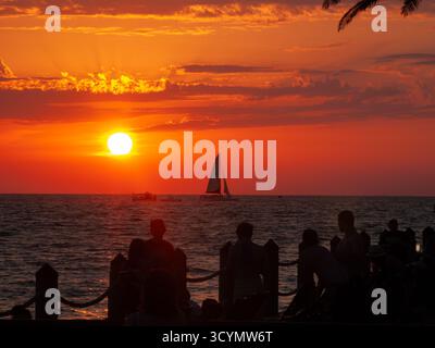 La gente guarda un tramonto vibrante sull'oceano, con una barca a vela sagomata contro il cielo ardente. Foto Stock