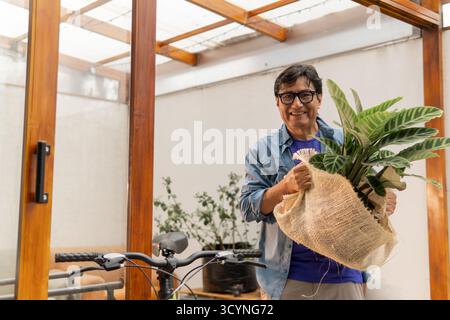 Uomo felice che porta la pianta in vaso avvolta a casa, godendo dell'hobby del giardinaggio Foto Stock