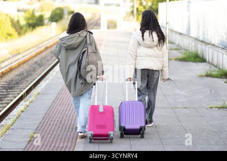 Due giovani amiche latine che camminano sulla piattaforma della stazione ferroviaria, tirando valigie colorate, preparandosi per un viaggio o arrivando da un viaggio Foto Stock