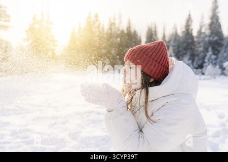 Una giovane donna vestita calorosamente con una giacca bianca e un cappello rosso a maglia si erge in una foresta innevata. Soffia allegramente dei fiocchi di neve nell'aria, creando Foto Stock