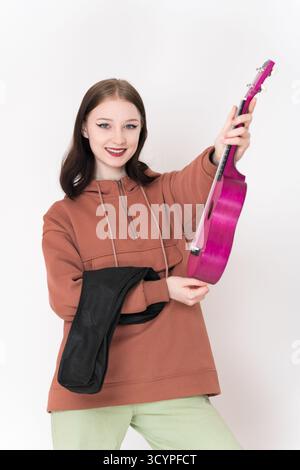 Una giovane donna positiva ukulelist tiene l'ukulele tra le mani e sorride guardando la telecamera. la donna di 18 anni indossa una felpa con cappuccio marrone e una jea verde chiaro Foto Stock