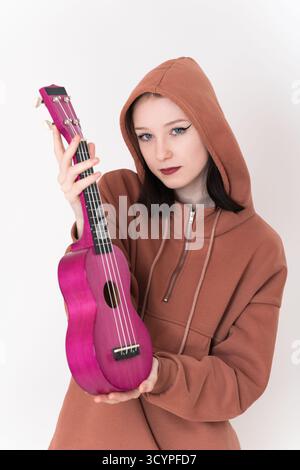Giovane donna giocatrice di ukulele che tiene l'ukulele in verticale tra le mani e guarda la fotocamera. Bella hipster caucasica con cappuccio marrone Foto Stock