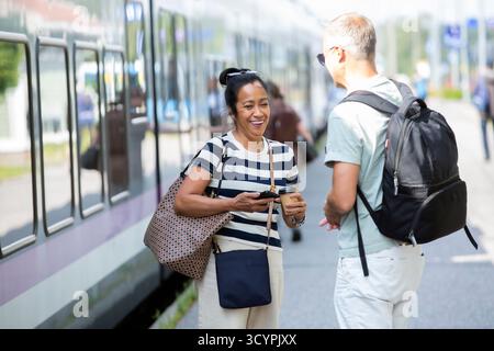 Donna asiatica felice di mezza età che ride con l'uomo sulla piattaforma del treno, simboleggia la diversità, la connessione, lo stile di vita moderno della città e gli spostamenti urbani. Foto Stock