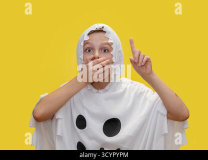 Il sorridente ragazzo vestito da fantasma punta verso l'alto, in piedi di fronte a un vibrante sfondo giallo, sorpreso di coprire con le mani la bocca per ha Foto Stock