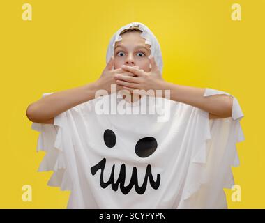 Il sorridente ragazzo vestito da fantasma punta verso l'alto, in piedi di fronte a un vibrante sfondo giallo, sorpreso di coprire con le mani la bocca per ha Foto Stock