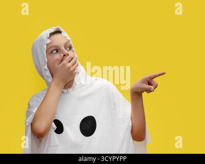 Il sorridente ragazzo vestito da fantasma punta verso l'alto, in piedi di fronte a un vibrante sfondo giallo, sorpreso di coprire con le mani la bocca per ha Foto Stock