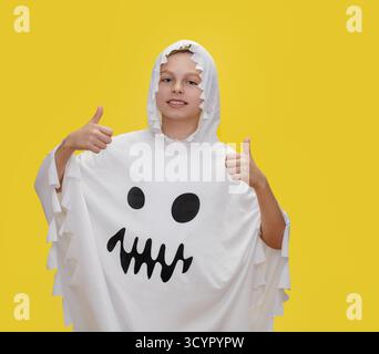 Il ragazzo sorridente vestito da fantasma punta verso l'alto, si trova davanti a uno sfondo giallo brillante e si alza il pollice, irradiando positività e sicurezza, l Foto Stock