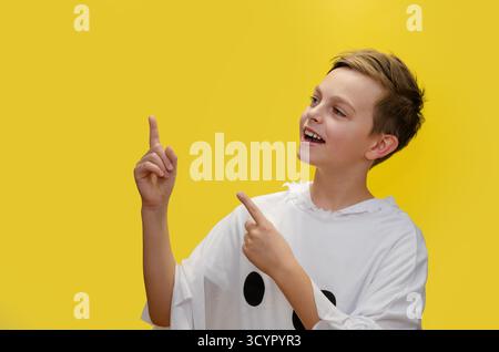 Il ragazzo sorridente vestito da fantasma punta verso l'alto, in piedi di fronte a un vivace sfondo giallo, probabilmente a tema per Halloween. Boy in Ghost Costume poi Foto Stock