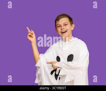 Il ragazzo sorridente vestito da fantasma punta verso l'alto, in piedi davanti allo sfondo viola, probabilmente a tema per Halloween. Ragazzo in costume fantasma che indica Upwar Foto Stock