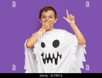 Il sorridente ragazzo vestito da fantasma punta verso l'alto, in piedi davanti a un vibrante sfondo viola, sorprende l'eccitazione di coprire con le mani la bocca per Hallo Foto Stock