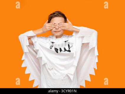 Il ragazzo sorridente vestito da fantasma punta verso l'alto, in piedi davanti allo sfondo arancione, probabilmente a tema per Halloween. Ragazzo in costume fantasma che indica Upwar Foto Stock