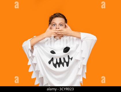 Il sorridente ragazzo vestito da fantasma punta verso l'alto, in piedi di fronte a un vivace sfondo arancione, sorpreso di coprire con le mani la bocca per ha Foto Stock