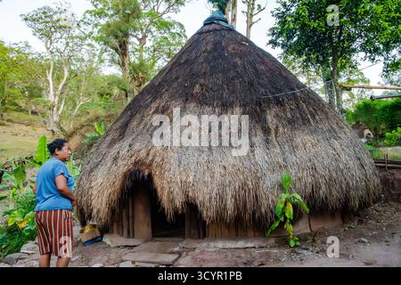 Una donna del posto in piedi di fronte alla sua tradizionale casa rotonda con tetto in paglia in erba sull'isola di Timor, Indonesia. Foto Stock