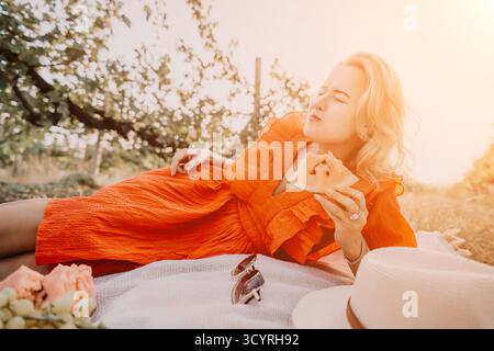Woman picnic Sunset Eating Food - una donna bionda in un abito arancione si gode un picnic in un frutteto al tramonto, mangiando una fetta di torta. Foto Stock