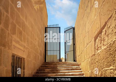 Una scalinata di pietra illuminata dal sole conduce ad aprire cancelli in ferro battuto tra le mura della vecchia fortezza, rivelando un cielo blu nuvoloso oltre. Foto Stock